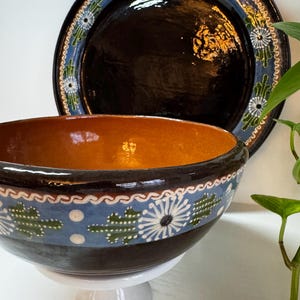 May include: A ceramic bowl and plate set. The bowl has a brown interior and a black exterior with a blue band featuring white floral and green cactus designs. The plate is black with a blue and white floral border.