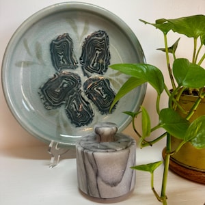 May include: A light blue ceramic plate with a floral design and five dark gray and white abstract shapes. A small, cylindrical marble jar with a lid sits in front of the plate. Green plant in a yellow pot.