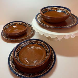 May include: A set of rustic, handmade ceramic bowls and plates. The bowls and plates are brown with a blue and white patterned rim. The set includes bowls and plates of different sizes, some stacked on a white cake stand.