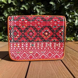 Könnte beinhalten: Rechteckige Tasche mit einem lebendigen roten und schwarzen geometrischen Muster, umrahmt von braunem Leder. Das Design der Tasche integriert verschiedene Farben. Sie wird auf einer Holzoberfläche präsentiert.