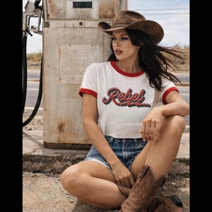 May include: A white ringer t-shirt with red trim on the sleeves and neckline, featuring the word Rebel in a red and black script font across the chest. The shirt is paired with blue denim shorts and a brown cowboy hat with a dark band.