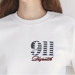 May include: A white crewneck sweatshirt featuring a graphic on the chest. The graphic displays the number 911 in a black and white striped pattern with a star field on the nine, and the word Dispatch written in a dark red cursive script below it.