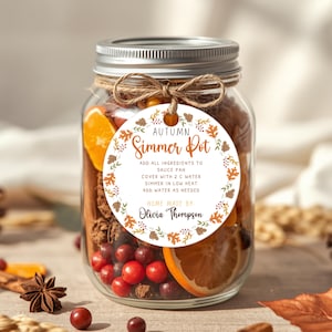 May include: A clear glass jar filled with dried orange slices, cranberries, and cinnamon sticks, topped with a silver lid and twine. A round white tag features a fall leaf border, the text Autumn Simmer Pot, instructions, and the name Olivia Thompson.