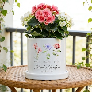 May include: A white ceramic Mom's Garden planter with a floral design and the names William, Alexander, and Emily. The planter holds pink and white flowers. The planter sits on a matching saucer on a wicker table.