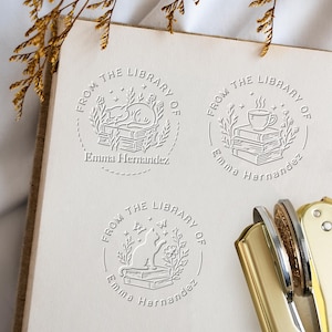 May include: A set of three embossed library stamps with the text "FROM THE LIBRARY OF" and the name "Emma Hernandez". Each stamp features a different design: a sleeping cat, a cup of coffee, and a cat with butterflies.