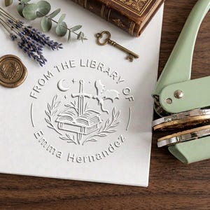 May include: A light green metal embosser tool next to a white paper featuring a circular embossed design. The design includes an open book with a sword, a crescent moon, stars, a dragon, and the text FROM THE LIBRARY OF Emma Hernandez.