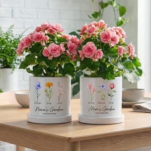 May include: Two white ceramic flower pots with pink flowers, each with a saucer. One pot reads "Nana's Garden" and the other reads "Mom's Garden". Both pots have floral illustrations and the text "Love Grows Here".