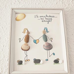 Puede incluir: Obra de arte enmarcada que presenta dos figuras hechas de piedras y conchas marinas, tomadas de la mano sobre un corazón. La obra incluye el texto "¡Te amo, Andrea... mi mejor amiga!" sobre un fondo blanco.