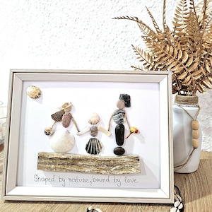Puede incluir: Obra de arte enmarcada que representa a una familia hecha de conchas y piedras. Las figuras se dan la mano, de pie sobre un trozo de madera flotante. El texto "Shaped by nature, bound by love" está escrito debajo. Un jarrón con hojas doradas en el fondo.