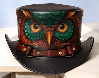 Puede incluir: Un sombrero de copa negro con una banda de cuero marrón y una ilustración detallada de un búho. El búho presenta ojos naranjas, detalles verdes y plumas marrones y naranjas. El sombrero tiene un ala curva.