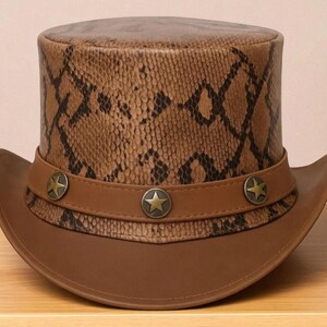 Puede incluir: Un sombrero de copa de cuero marrón con un estampado de piel de serpiente en la corona. El sombrero presenta un ala ancha y una banda de cuero marrón con tres adornos metálicos en forma de estrella. Un accesorio elegante.
