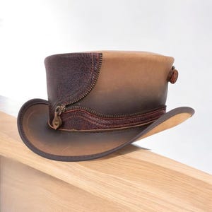 Puede incluir: Un sombrero de copa de cuero marrón con un detalle de cremallera. El sombrero presenta un ala ancha y un diseño de dos tonos, con una banda y detalles en marrón oscuro. El sombrero mide aproximadamente 15 cm de alto.