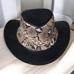May include: A black cowboy hat with a snakeskin pattern band. The hat has a wide brim and a rounded crown. The snakeskin band is tan, brown, and white. The hat is made of leather.