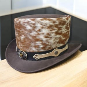 May include: A brown cowboy hat with a unique design. The hat features a brown and white spotted fur crown, a dark brown brim, and a black band with decorative metal accents. The hat has a classic western style.