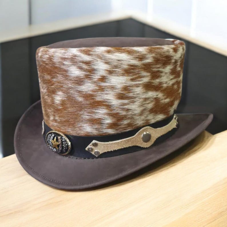 May include: A brown cowboy hat with a unique design. The hat features a brown and white spotted fur crown, a dark brown brim, and a black band with decorative metal accents. The hat has a classic western style.