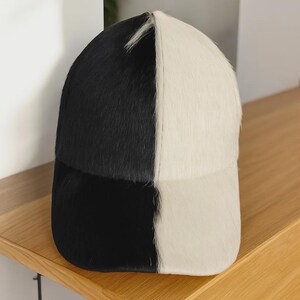 Puede incluir: Gorra de béisbol bicolor en blanco y negro. La gorra está dividida verticalmente, con un lado negro y el otro blanco. El material tiene un aspecto suave y texturizado, posiblemente piel o un tejido similar. Un accesorio elegante.