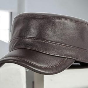 Puede incluir: Gorra de estilo militar de cuero marrón oscuro con una visera corta. La gorra presenta una corona estructurada y un detalle de banda sobre la visera. El cuero tiene un aspecto ligeramente texturizado y las costuras son visibles.