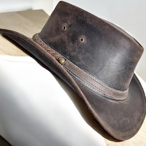 May include: A distressed brown leather hat with a wide brim and a tall crown. The hat features a decorative band around the crown and a braided detail. The hat has two small air vents on the crown and a metal button detail.