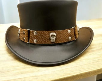 May include: A black leather top hat with a brown snakeskin band featuring silver skull accents and rivets. The hat has a wide brim and a tall, cylindrical crown. The hat is displayed on a light-colored wooden surface.