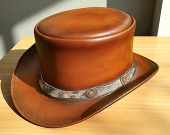 May include: A brown leather top hat with a wide brim. The hat features a decorative band with circular accents. The hat is made of a smooth, shiny leather and sits on a wooden surface.