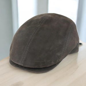 May include: A brown suede flat cap with a rounded crown and a small, stiff peak. The cap has a stitched seam down the center and is made of a soft, textured material. A classic hat style.