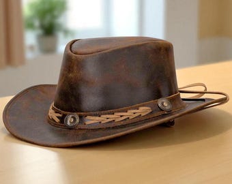 May include: A brown leather cowboy hat with a distressed finish. The hat features a decorative band with stitched leather accents and two round metal studs. The brim is wide, and the crown is rounded.