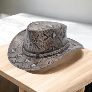 Può includere: Un cappello da cowboy con motivo a pelle di serpente nei toni del marrone, beige e grigio. Il cappello presenta una fascia in pelle marrone scuro con cuciture decorative e un bordo intrecciato. Il cappello è in pelle.