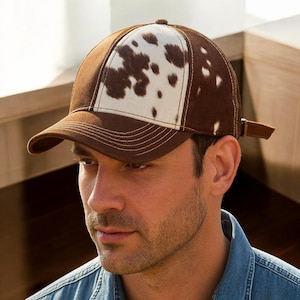 Puede incluir: Gorra de béisbol marrón y blanca con estampado de piel de vaca. La gorra tiene una visera marrón y un panel blanco con manchas marrones. Con correa ajustable marrón y costuras blancas.