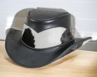 May include: Black leather cowboy hat with a cowhide band featuring black and white patches. The hat has a braided band around the crown and a wide brim. The hat is displayed on a wooden surface.