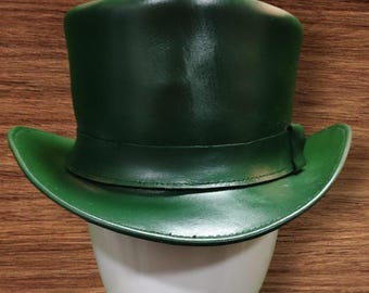 Puede incluir: Un sombrero de copa verde oscuro con ala ancha y una banda alrededor de la corona. El sombrero está hecho de un material liso. El sombrero se exhibe sobre una cabeza de maniquí blanca sobre un fondo de vetas de madera.