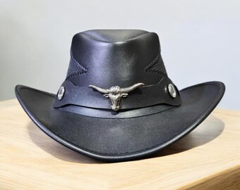 May include: A black cowboy hat with a silver bull head emblem on the front. The hat has a wide brim and a structured crown. The hat is made of a leather-like material and has decorative stitching.