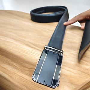 May include: A black leather belt with a silver buckle. The buckle is rectangular with a glossy black finish and silver detailing. The belt rests on a light brown wooden surface.