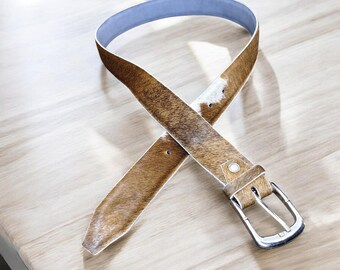 May include: A brown and white cowhide belt with a silver buckle and a light blue interior. The belt is crossed over itself, showcasing the texture and pattern of the leather. The buckle is rectangular.