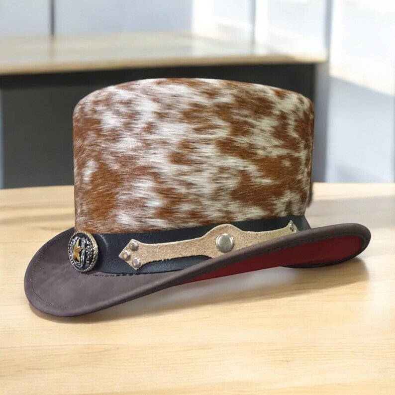 May include: A brown and white cowhide top hat with a wide brim. The hat features a black band with decorative metal accents and a star-shaped emblem. The underside of the brim is red.