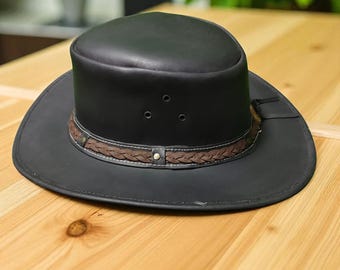 May include: A black leather cowboy hat with a wide brim and a brown braided band. The hat has three small air vents on the crown and a leather strap detail. The hat is sitting on a wooden surface.