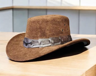 May include: A brown suede hat with a wide brim and a decorative band. The hat features a leather band in shades of gray and silver. The hat has a classic, outdoorsy style.