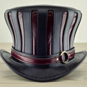 Puede incluir: Un sombrero de copa de cuero negro con detalles burdeos. El sombrero presenta paneles burdeos verticales entre paneles negros, una banda burdeos con una hebilla dorada y un ala ancha. Hecho a mano.
