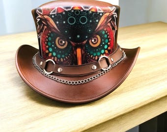 Puede incluir: Sombrero de copa marrón con ala ancha y diseño decorativo de búho. El sombrero presenta una banda de cuero marrón con cadena y remaches plateados. El diseño del búho incluye colores rojo, verde y naranja.