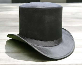 May include: A black top hat with a wide brim and a dark gray ribbon band. The hat is made of a smooth, possibly velvet-like material. The hat is sitting on a light-colored wooden surface.