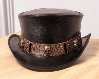 May include: Black leather top hat with a decorative band featuring a snakeskin pattern and skull accents. The hat has a classic top hat shape with a rounded crown and a curved brim. The band is adorned with metal skull embellishments.