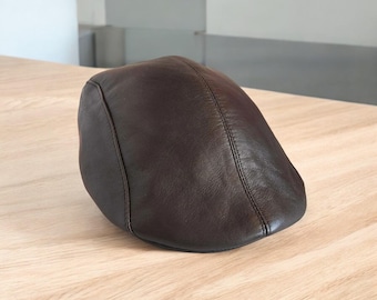 May include: A brown leather flat cap with a rounded crown and a small, stiff brim. The cap is made of a smooth, dark brown leather and has a stitched seam detail. The cap is sitting on a light-colored wooden surface.