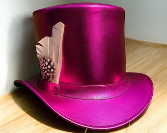 Op de afbeelding: Een magenta hoge hoed met een brede rand en een decoratieve veer. De hoed heeft een glanzende, metallic afwerking en een band rond de kroon. De veer is lichtbruin met donkerbruine vlekken.