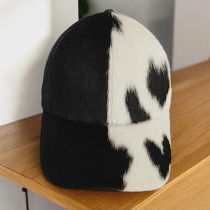 May include: A black and white cowhide baseball cap. The cap is split down the middle, with one half black and the other half white with black spots. The cap is sitting on a wooden surface.