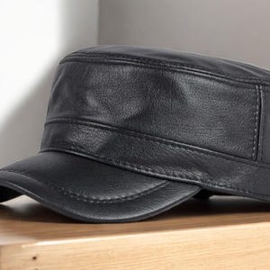 Pode incluir: Boné militar de couro preto com parte superior plana e aba curta e curva. O boné tem um design estruturado com costuras visíveis e um acabamento liso e fosco. O boné está sobre uma superfície de madeira.