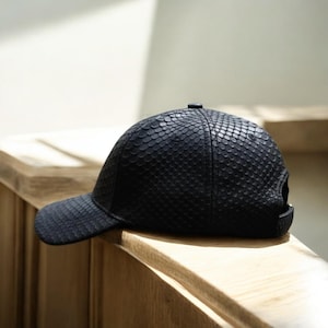 Könnte beinhalten: Schwarze Baseballkappe mit einem strukturierten, schuppenartigen Muster. Die Kappe hat einen gebogenen Schirm und einen verstellbaren Riemen am Rücken. Die Kappe liegt auf einer Holzoberfläche.