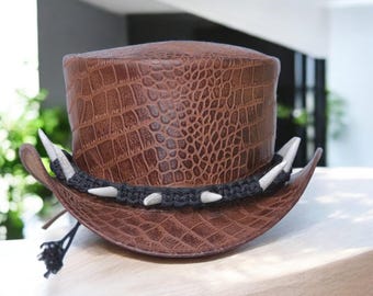 Peut inclure: Chapeau haut de forme en cuir marron avec une texture peau de crocodile. Le chapeau est orné d'une bande noire avec des pointes blanches et un petit pompon noir. Il a un large bord et une haute calotte.
