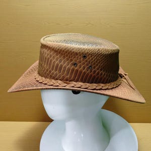 Puede incluir: Un sombrero de vaquero color canela con un patrón texturizado similar a la piel de serpiente en la copa y el ala. El sombrero incluye una banda de cuero trenzado en la base de la copa y tres pequeños ojales de ventilación de metal oscuro.