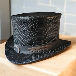 Peut inclure: Un chapeau haut de forme noir avec une texture peau de serpent. Le chapeau a un large bord et une bande noire avec un motif décoratif en cuir marron. Le chapeau est fait d'un matériau texturé.