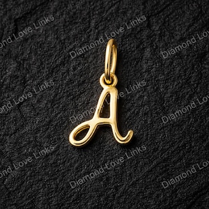 Peut inclure: Un pendentif doré en forme de la lettre "A" en écriture cursive. Le pendentif a une petite boucle en haut pour une chaîne. Le fond est une surface noire texturée.