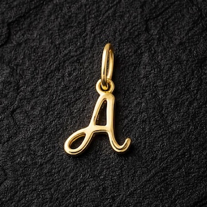 Puede incluir: Un colgante dorado con la forma de la letra "A" en escritura cursiva. El colgante tiene un pequeño lazo en la parte superior para sujetarlo a una cadena. El fondo es una superficie negra texturizada.